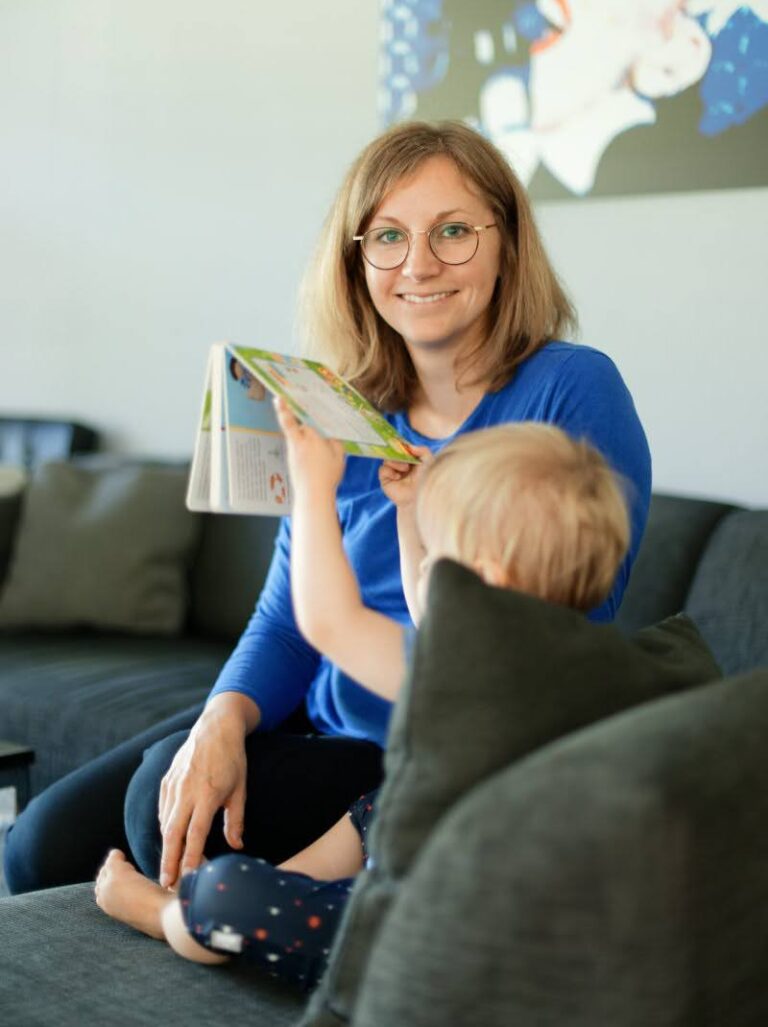 Caucasian woman sitting on a sofa smiling and looking at the camera above the head of a toddler sitting on the sofa who's holding up a book