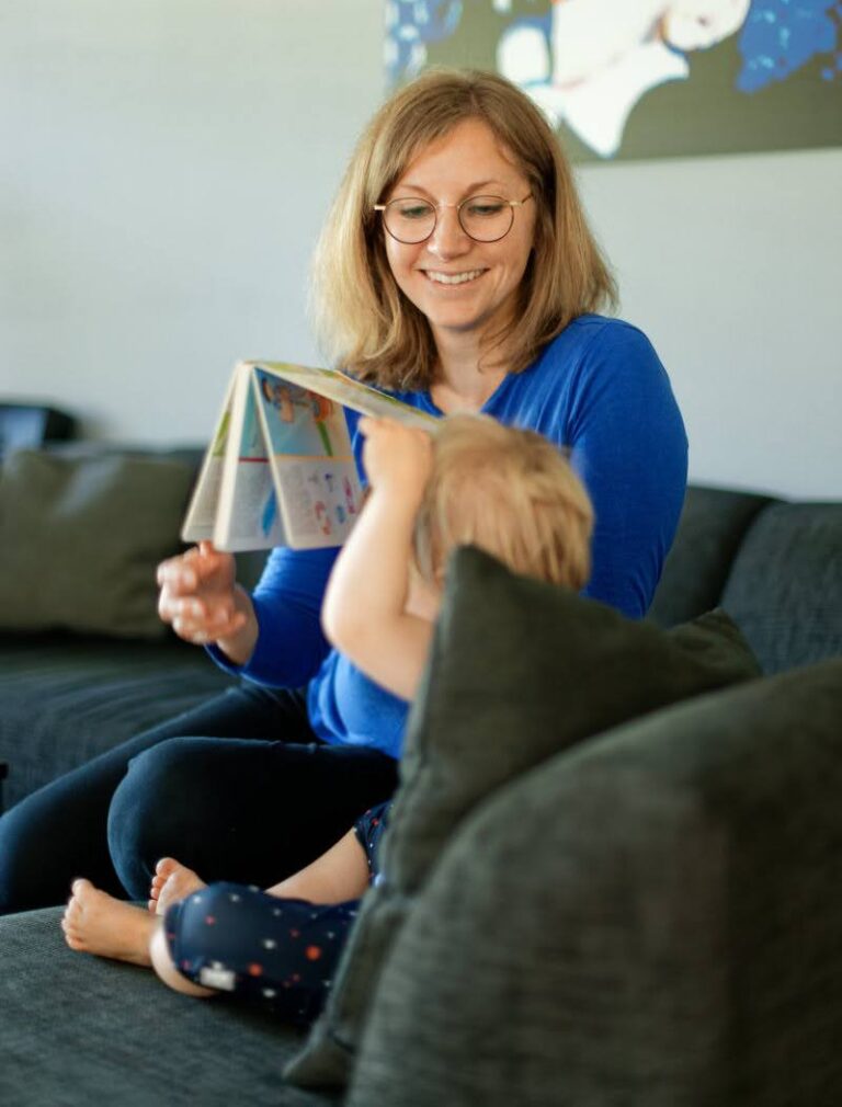 Caucasian woman sitting on a sofa smiling at a toddler who's holding up a book