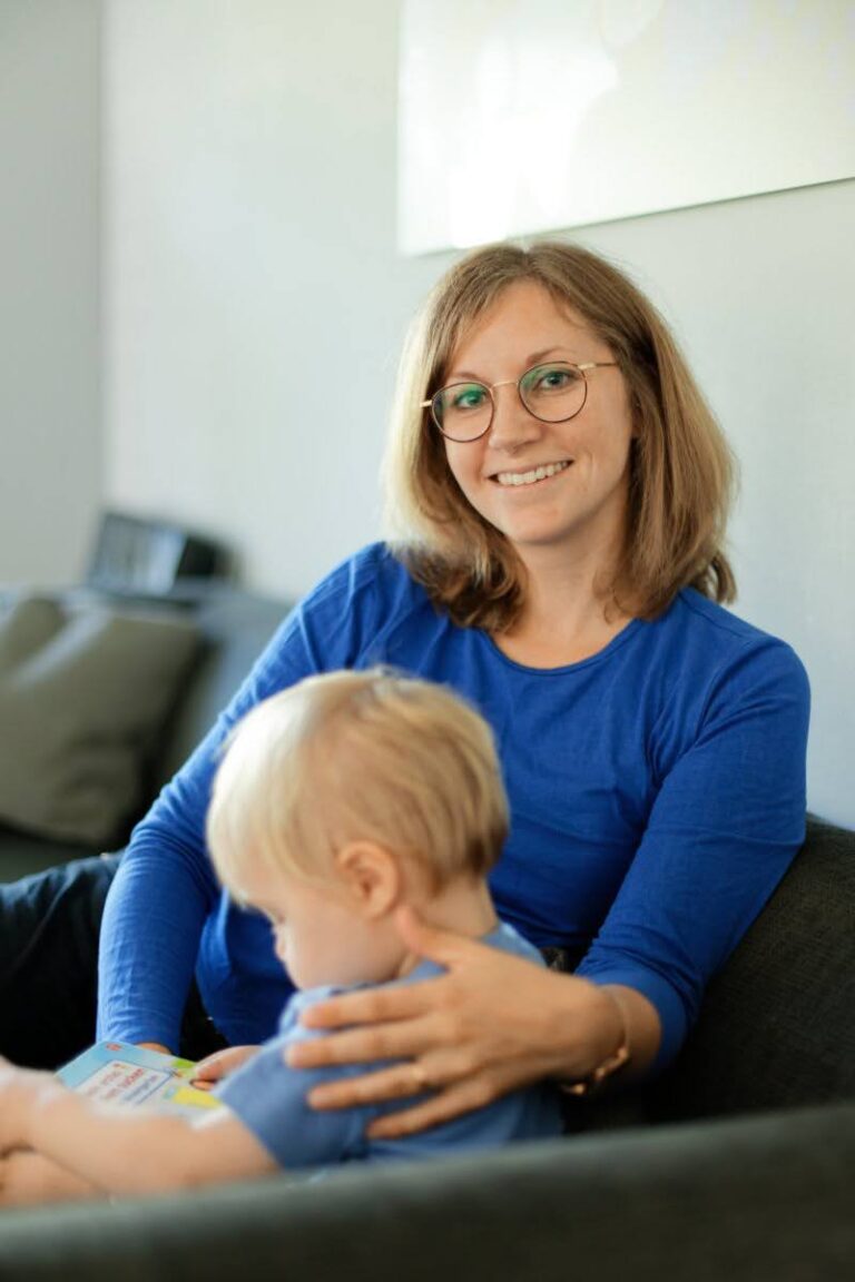 Caucasian woman sitting on a sofa with her arm around a toddler while smiling at the camera
