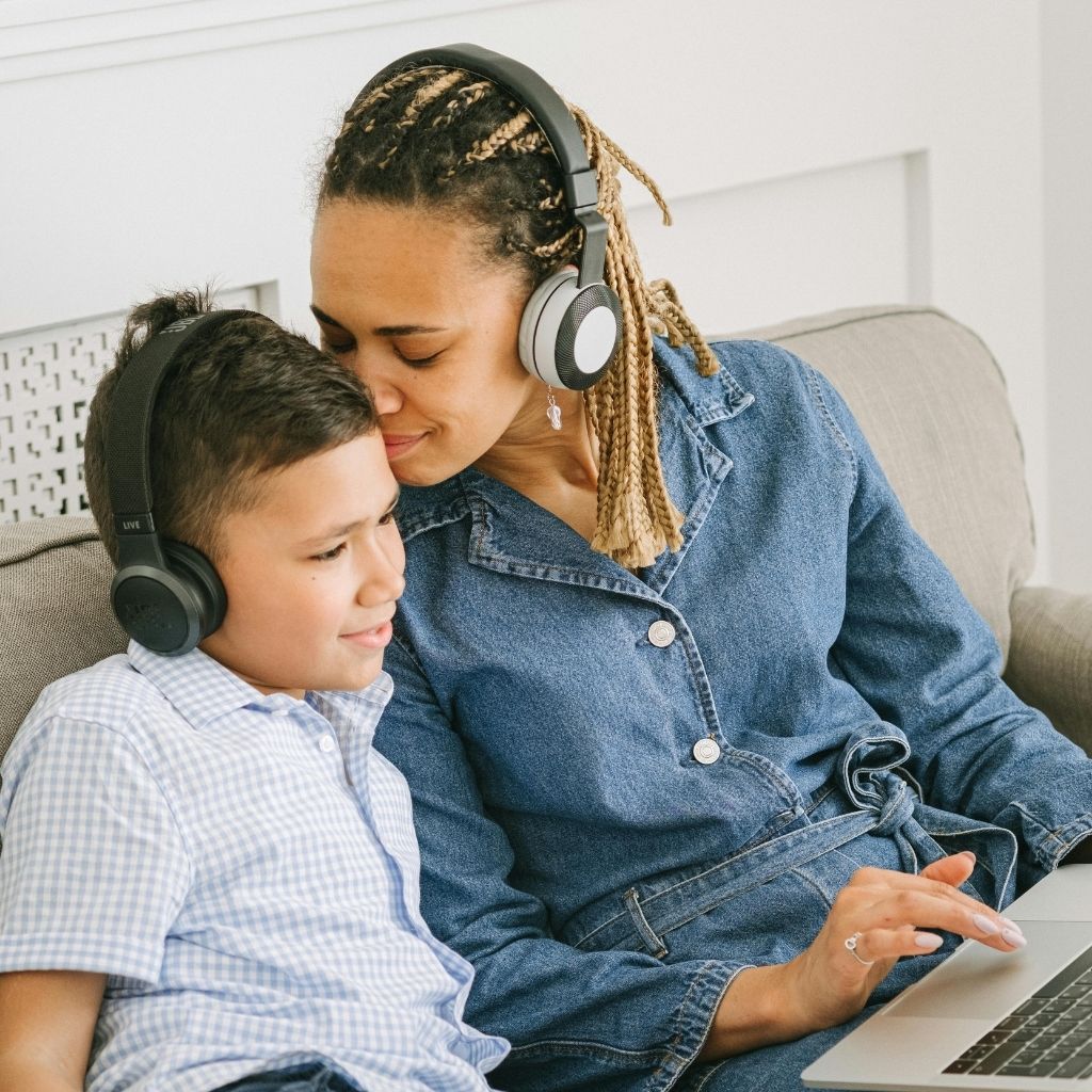 Woman with headphones kissing a boy on the head who's sitting next to her on the sofa and is looking at a laptop wearing headphones