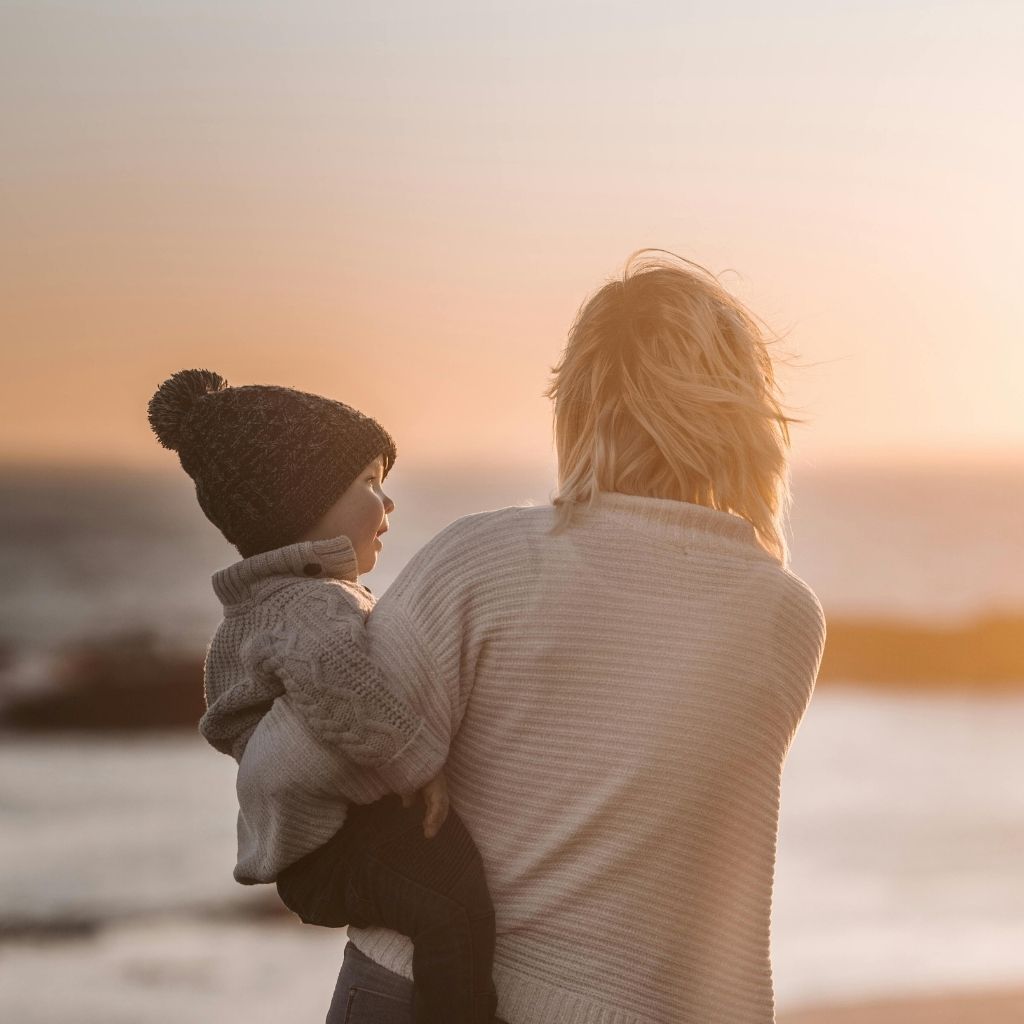 Caucasian woman holding a toddler while looking at the sea in sunset